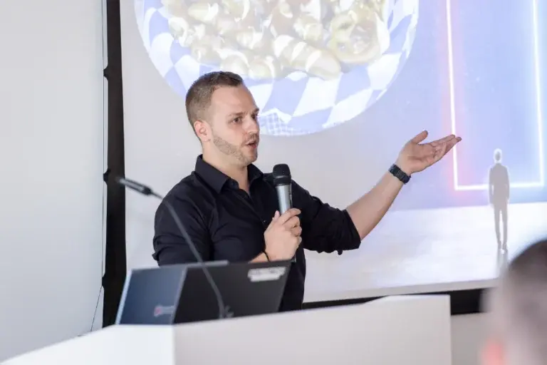 Ein Vortragender spricht mit Mikrofon vor einer Projektion und gestikuliert zum Publikum.