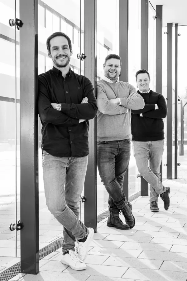Florian Dodegge, Gerald Eder und Manuel Dorfer lehnen mit verschränkten Armen hintereinander an den Glassäulen eines hellen Gangs.