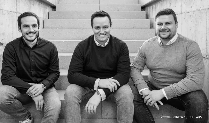 Florian Dodegge, Manuel Dorfer und Gerald Eder sitzen nebeneinander auf einer Betontreppe und lächeln in die Kamera.