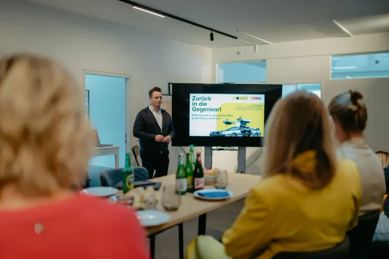 Manuel Dorfer spricht vor einer Gruppe an einem gedeckten Tisch, neben ihm ein Bildschirm mit einer Präsentation.